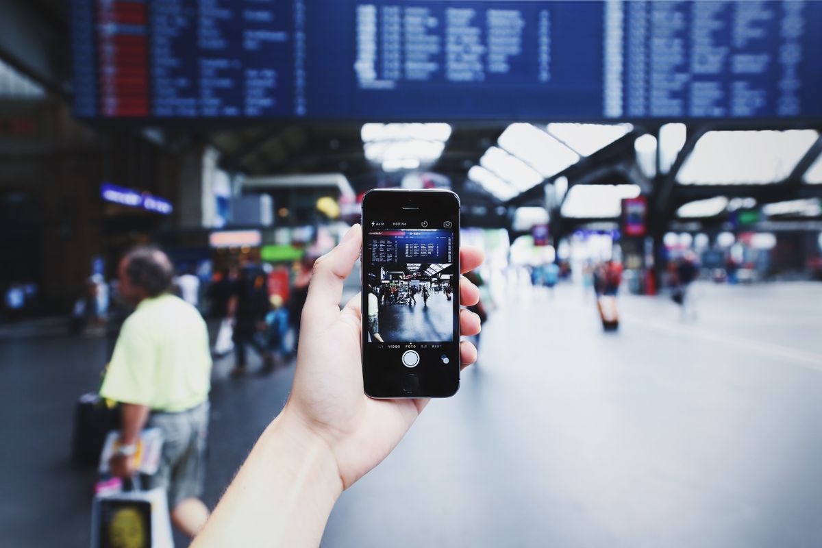 Mano che tiene uno smartphone in una stazione affollata con il tabellone delle partenze sullo sfondo