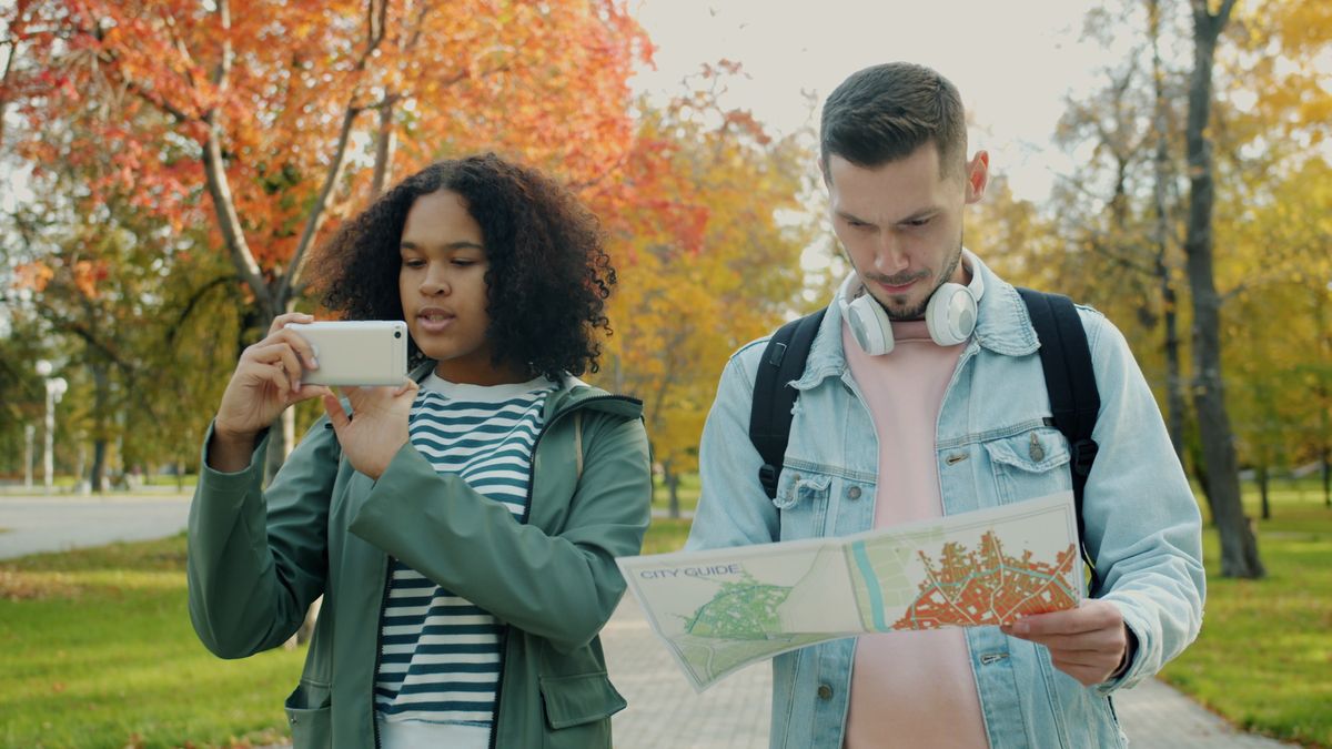 Dos jóvenes viajeros en un parque, uno usando el teléfono mientras el otro mira un mapa de la ciudad