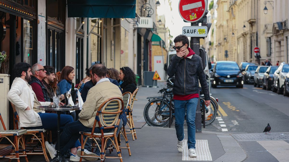 Gente en la terraza de un café de París mientras un hombre pasa mirando su teléfono