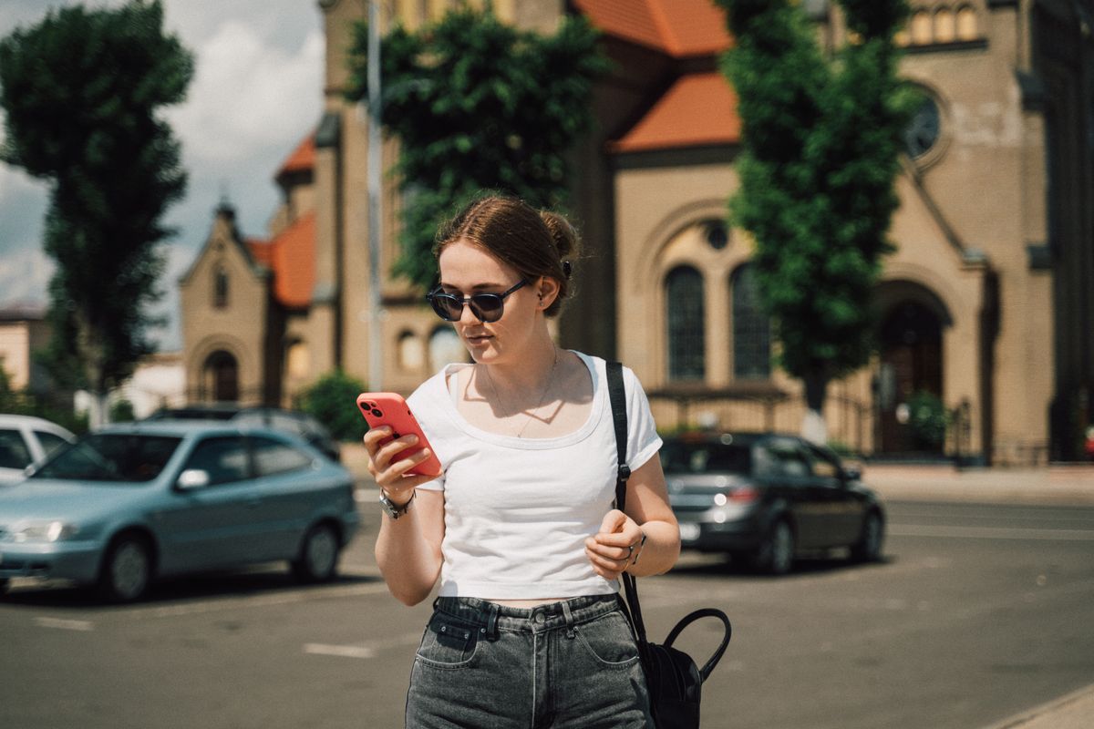 Mujer joven con lentes de sol revisando su smartphone en una calle soleada de un pueblo antiguo de Europa